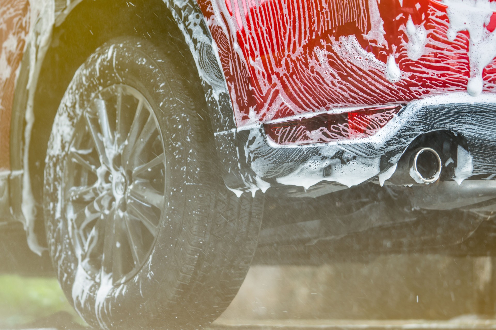 Red compact SUV car with sport and modern design washing with soap. Car covered with white foam. Car care service business concept. Car wash with foam before glass waxing and glass coating automobile.